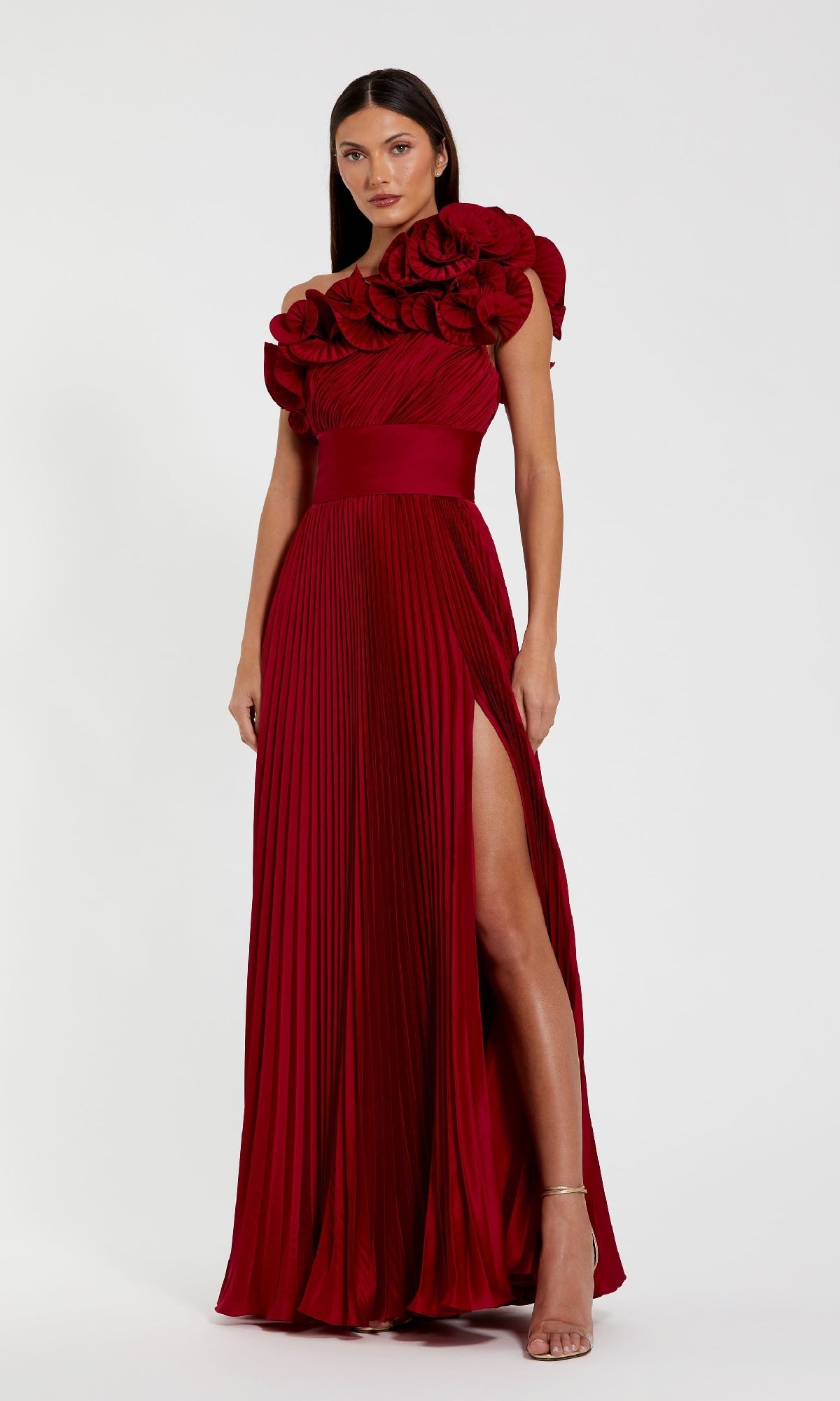 A woman models the Mac Duggal 49902 Pleated One-Shoulder Prom Gown, featuring oversized floral ruffles on one shoulder and a high slit, against a plain white background.